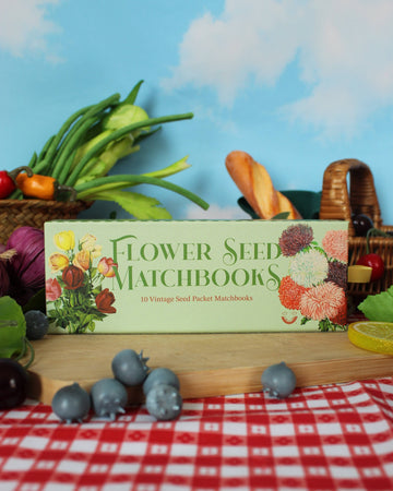 Box of Flower Seed Matchbooks on a table with vegetables and a picnic basket in the background.