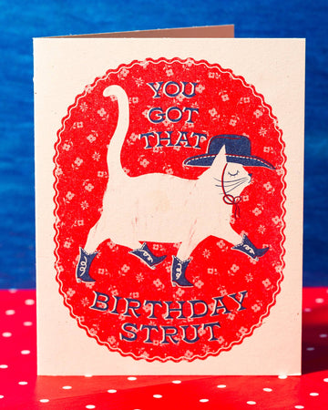 A folded risograph card with a red and blue background, featuring an illustration of a cat wearing boots and the text 'You GOT THAT BIRTHDAY STRUT' on the front, with a polka dot pattern on the back.