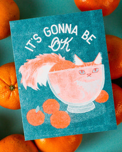A risograph card with a cartoon image of a cat inside a bowl, with the text 'It's Gonna Be OK' in a circular pink background, surrounded by orange and teal colors.