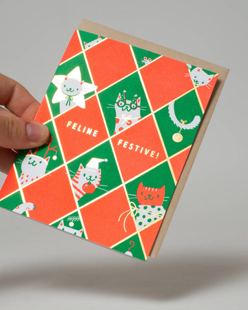 Greeting card with cat illustrations and 'Feline Festive!' text held by a hand on a gray background