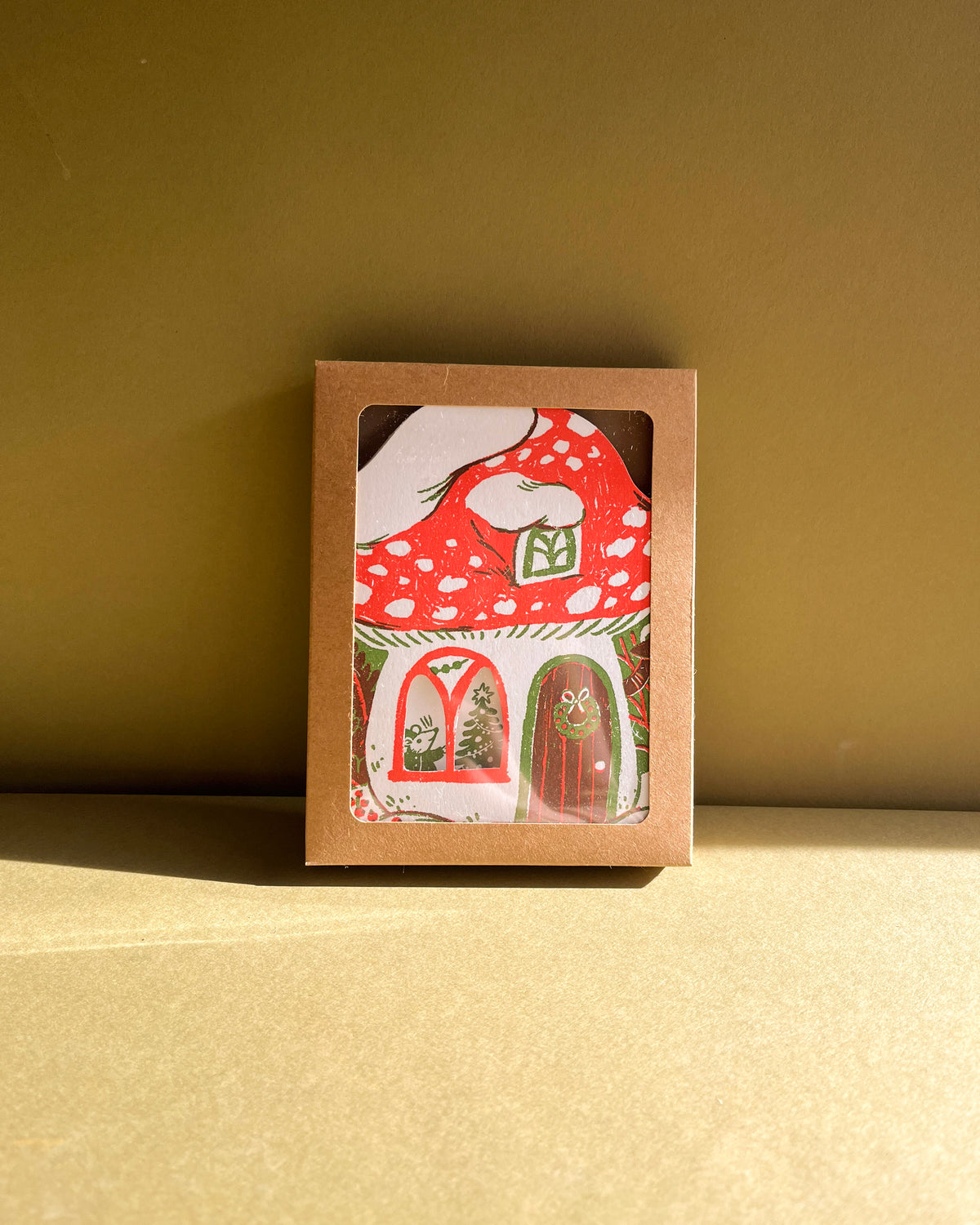 Card with a red mushroom house design in a brown box on a beige surface.