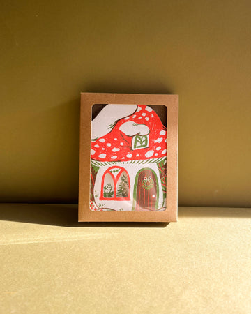 Card with a red mushroom house design in a brown box on a beige surface.