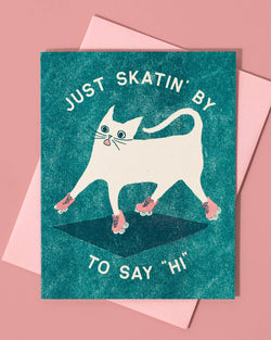A folding greeting card with a illustration of a cat wearing rollerskates on a teal and light mauve background. The text on the card reads 'JUST SKATIN' BY TO SAY ‘HI’.