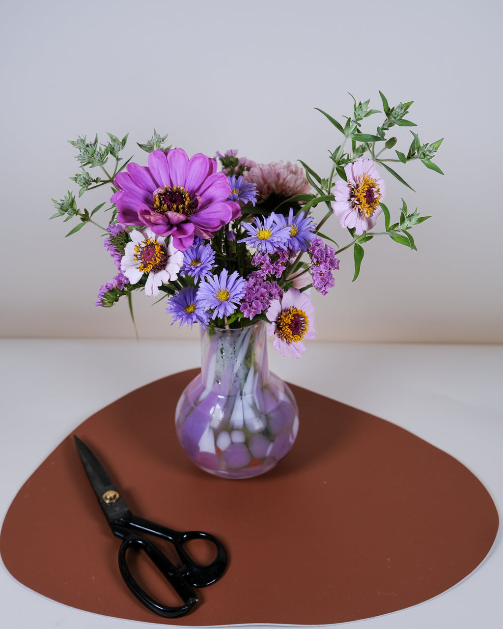 A small clear glass vase filled with a mixed flower bouquet in purple, pink, white, and green, placed on a brown oval mat with black-handled scissors in front, against a plain light background.