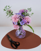 A bouquet of pink and purple flowers arranged in a small clear glass vase, placed on a brown oval mat with black-handled scissors beside it against a plain light background.