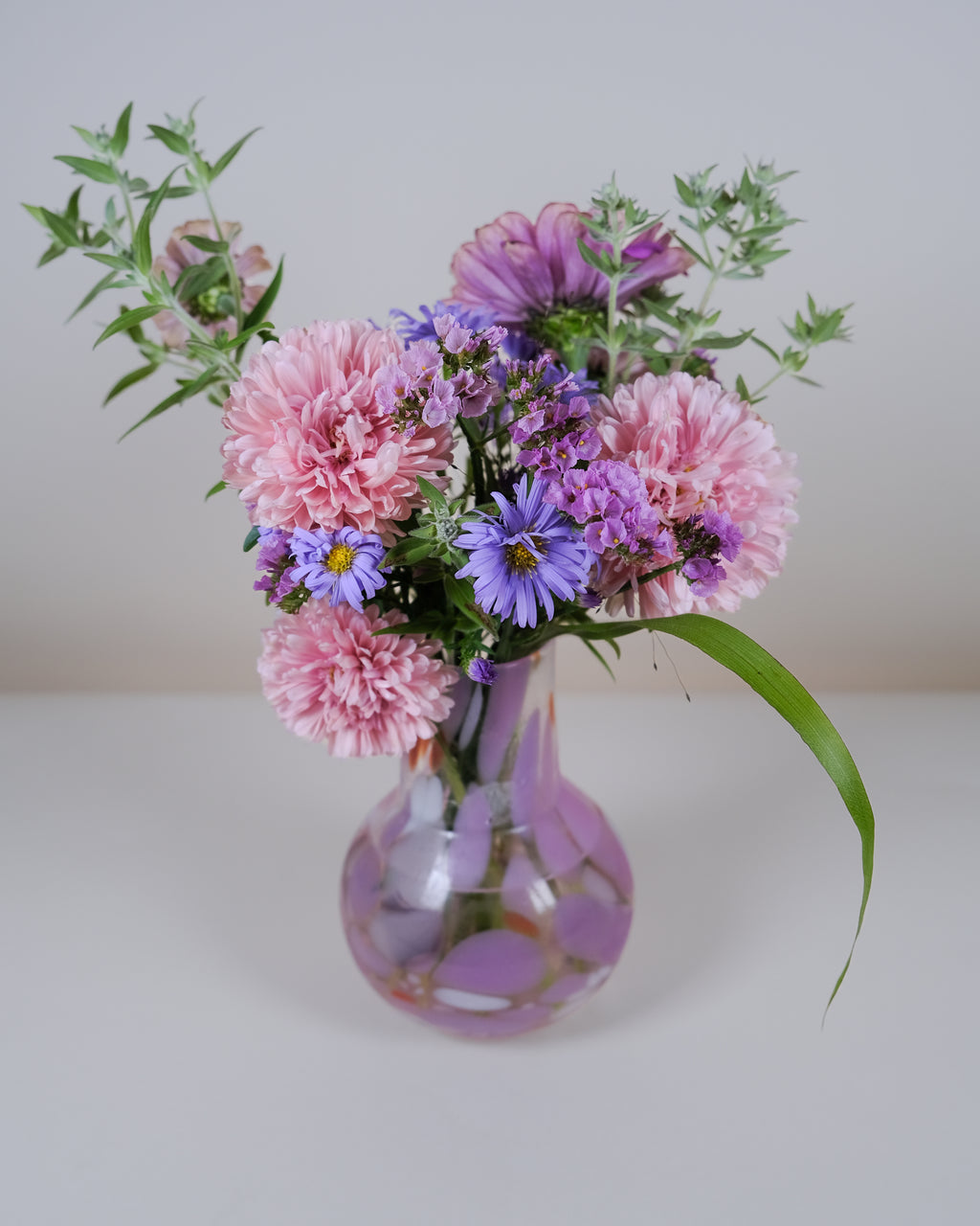 A floral arrangement of pink carnations, purple asters, and green foliage in a clear glass vase with a purple pattern, set against a plain light gray background.