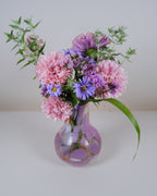 A floral arrangement of pink carnations, purple asters, and green foliage in a clear glass vase with a purple pattern, set against a plain light gray background.