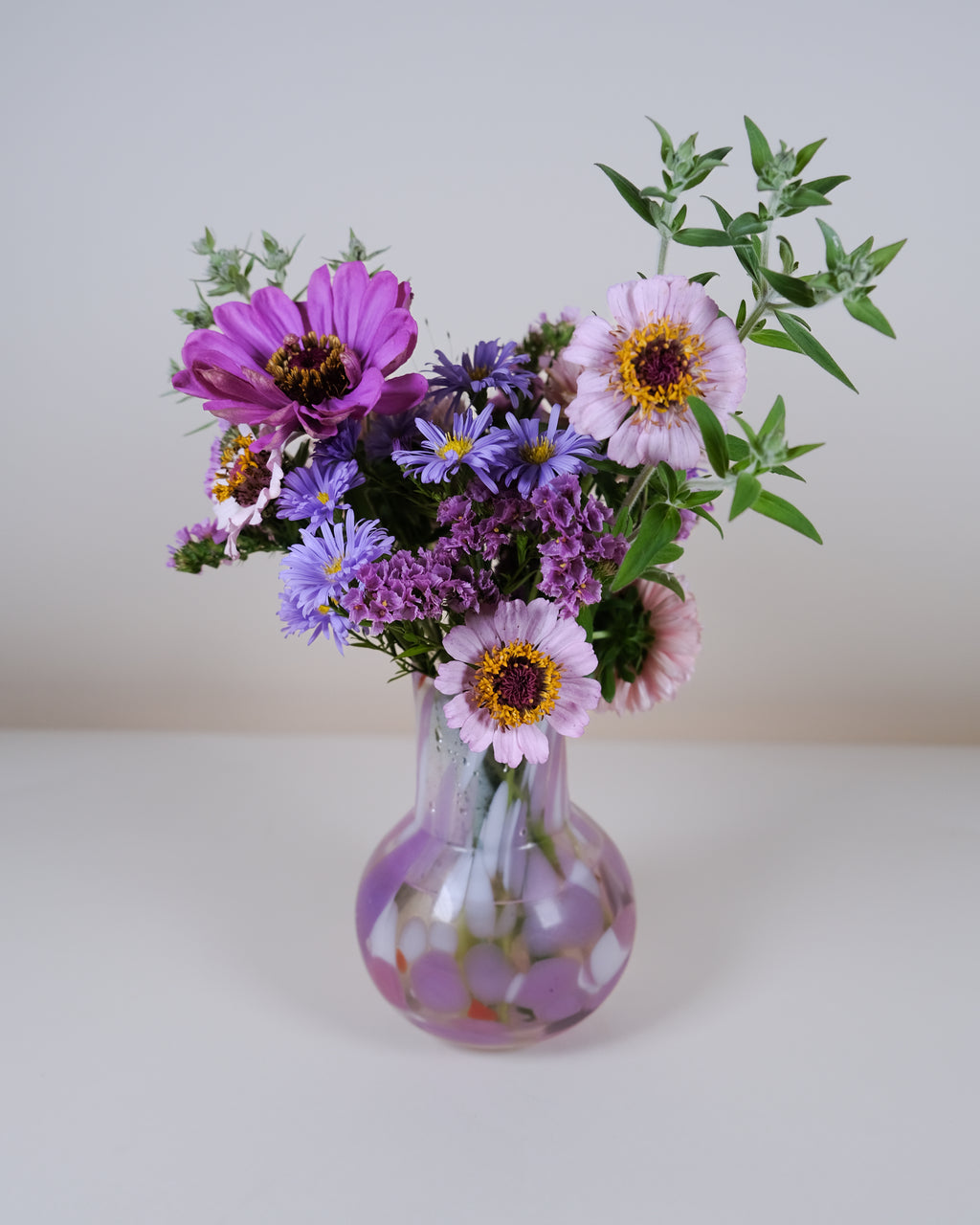 A mixed flower bouquet in a small clear glass vase with a rounded bulb shape. The bouquet features purple and pink flowers with green foliage, set against a plain light gray background.