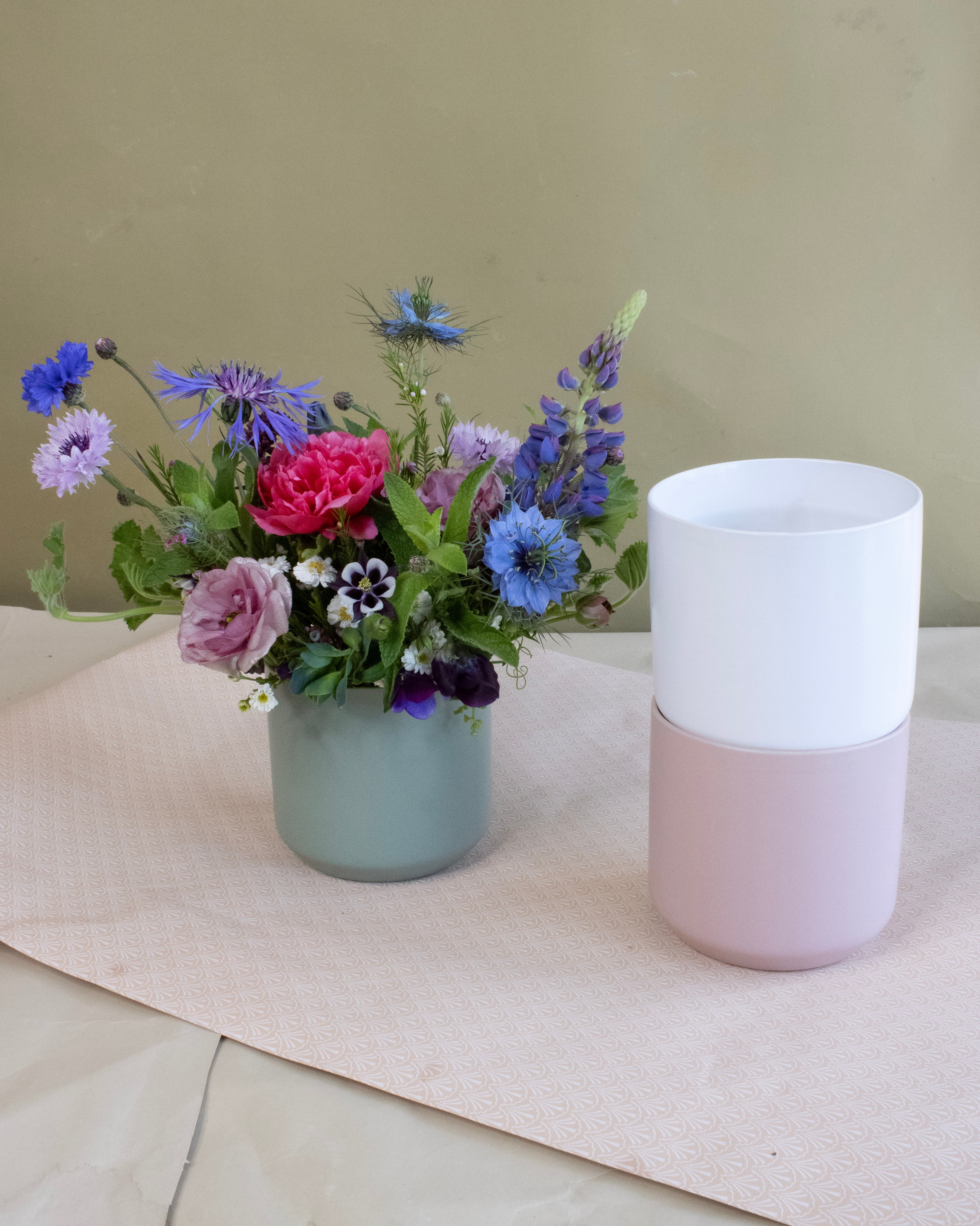 Bouquet of flowers in a green vase next to pink and white vases on a light surface.