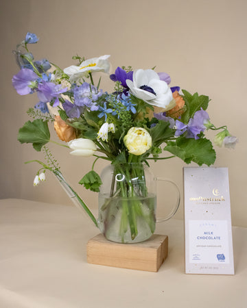 Arrangement of flowers in a clear vase on a wooden block with a bar of milk chocolate next to it.