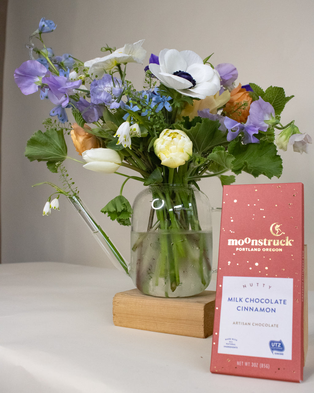 Arrangement of flowers in a vase next to a bar of moonstruck chocolate on a white surface.
