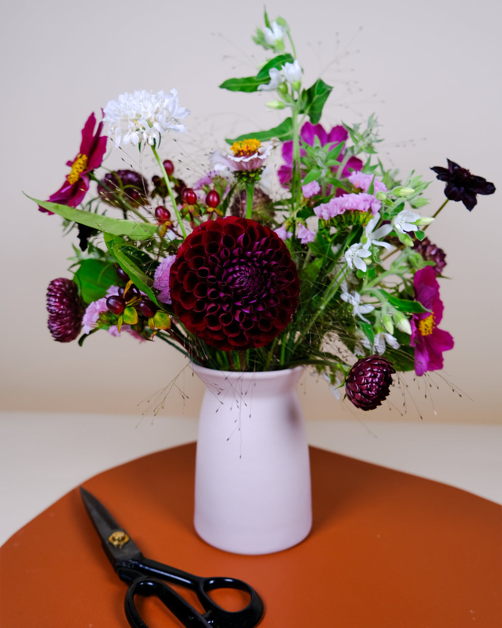 A mixed floral arrangement in a pale pink ceramic vase, placed on an orange surface against a beige background. A pair of black-handled scissors rests beside the vase.