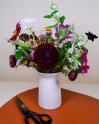 A mixed floral arrangement in a pale pink ceramic vase, placed on an orange surface against a beige background. A pair of black-handled scissors rests beside the vase.