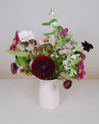 A mixed wildflower arrangement in a pale pink ceramic pitcher vase, featuring burgundy, magenta, white, and purple flowers with green foliage, set against a neutral light gray background.