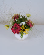 A floral arrangement featuring red and yellow flowers with green foliage, styled in a small white ceramic vase against a plain light gray background.