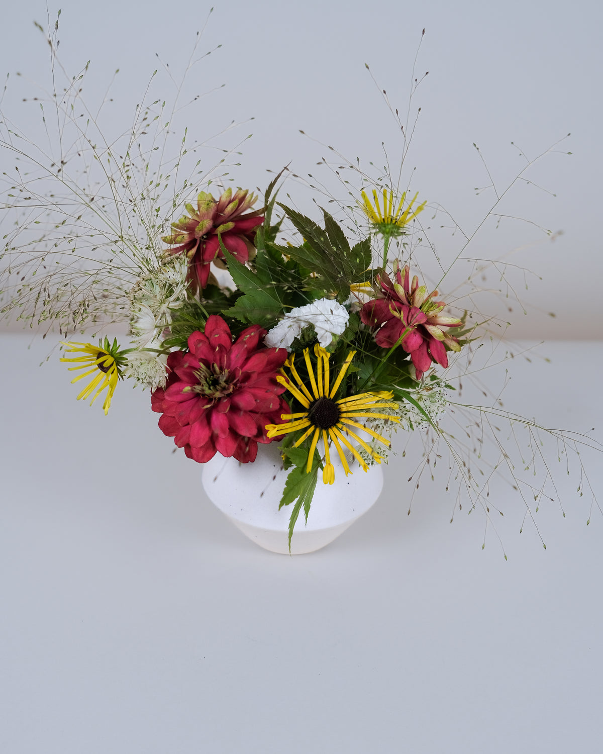 A floral arrangement featuring red and yellow flowers with green foliage, styled in a small white ceramic vase against a plain light gray background.