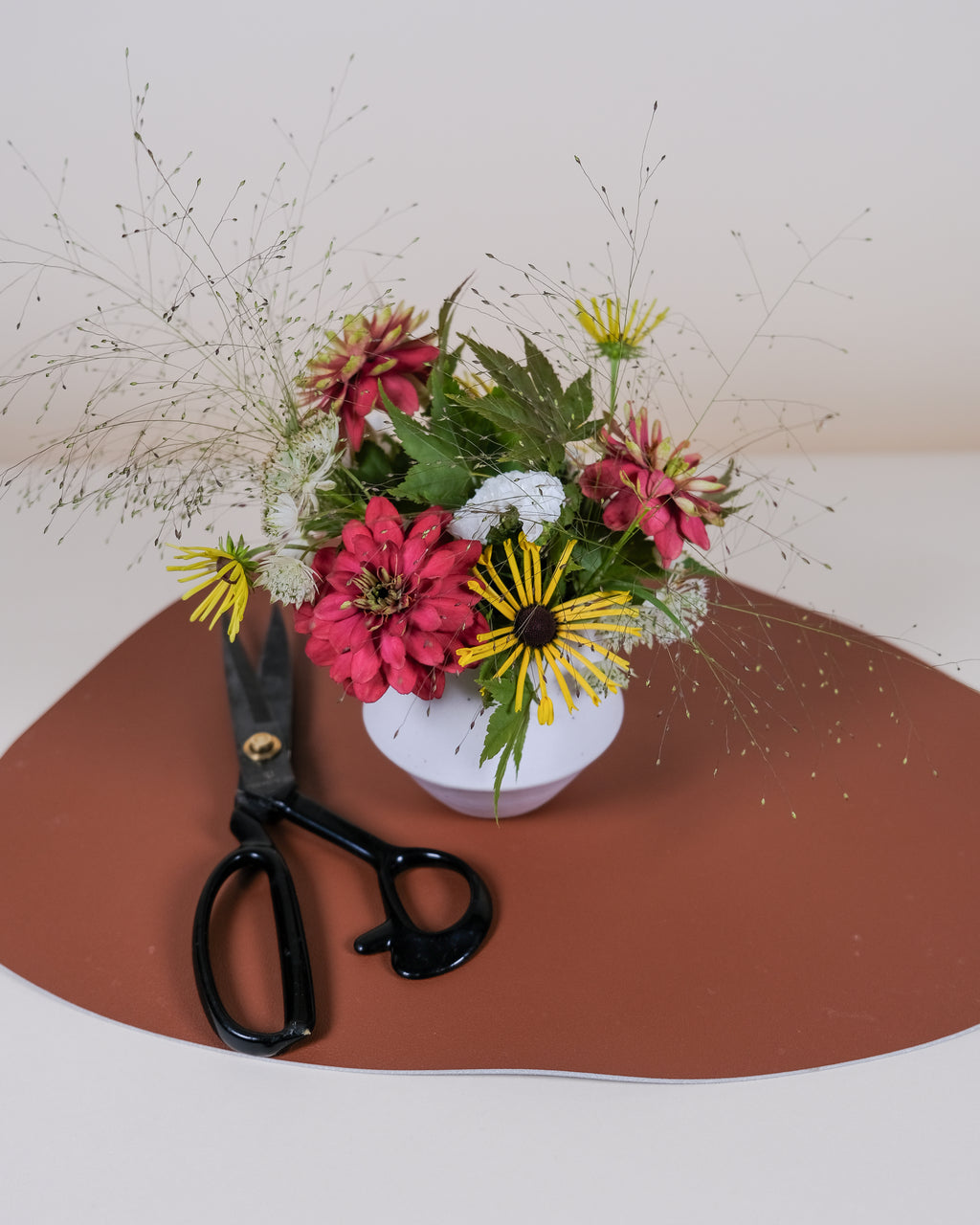 A small floral arrangement in a white vase with pink, yellow, and green flowers, placed on a terracotta-colored surface with black-handled scissors beside it against a plain light background.