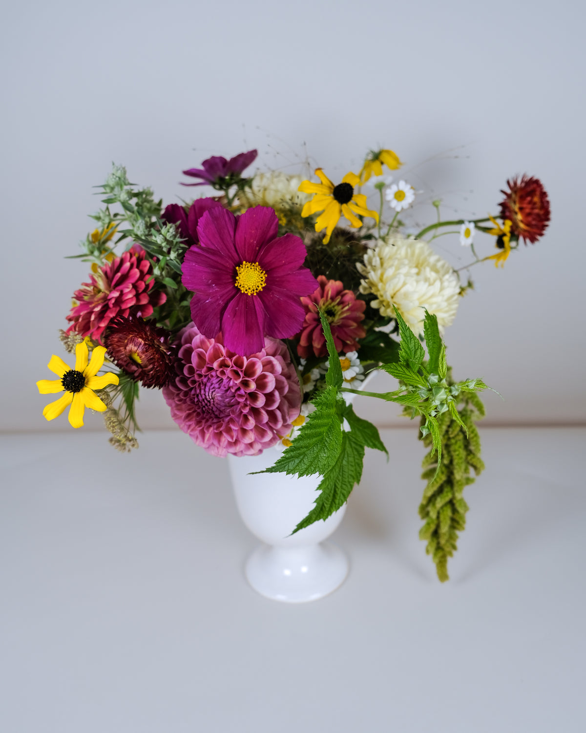 A vibrant mixed flower bouquet in a white ceramic vase, featuring a large magenta cosmos, pink chrysanthemums, yellow black-eyed Susans, white daisies, and green foliage, set against a plain light gray background.