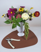 A white ceramic vase filled with a colorful wildflower arrangement, placed on a brown irregular-shaped mat with pruning shears on a white surface against a light gray background.