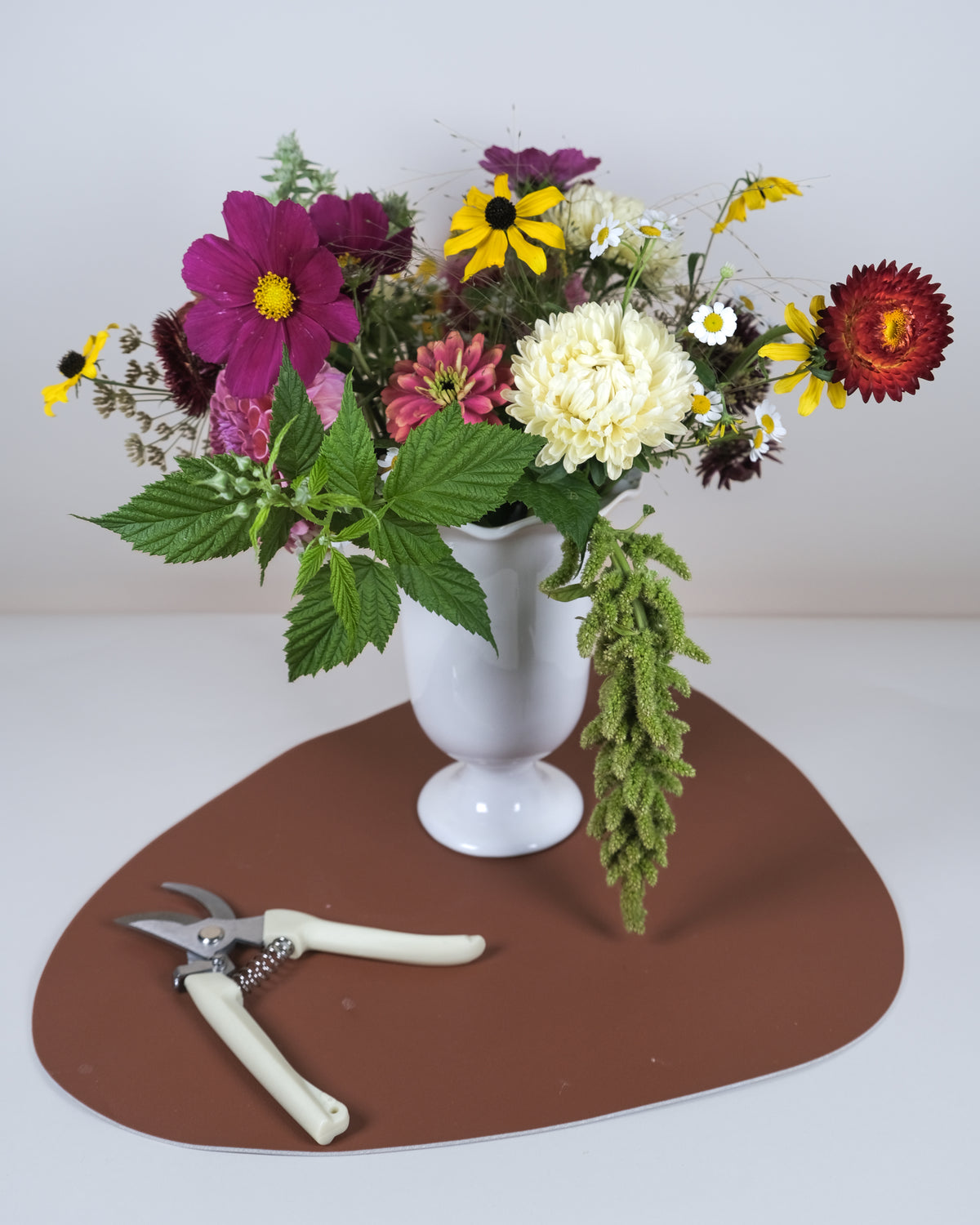 A white ceramic vase filled with a colorful wildflower arrangement, placed on a brown irregular-shaped mat with pruning shears on a white surface against a light gray background.