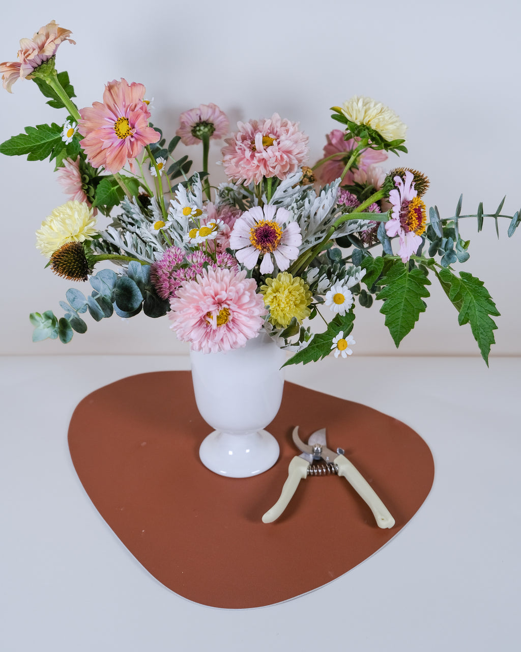 A colorful wildflower bouquet in a white ceramic vase, displayed on a brown heart-shaped mat with a pair of pruning shears, set against a plain white background.