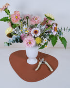 A colorful wildflower bouquet in a white ceramic vase, displayed on a brown heart-shaped mat with a pair of pruning shears, set against a plain white background.