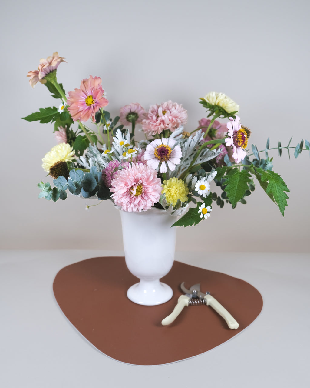 A floral arrangement of pink, yellow, white, and green flowers in a white pedestal vase, placed on a brown heart-shaped mat with pruning shears nearby against a plain light gray background.