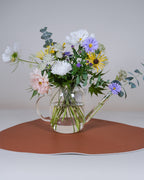 A floral arrangement in a clear glass teapot vase, featuring white, purple, yellow, and pink flowers with green stems and eucalyptus leaves, set on a terracotta-colored mat against a neutral gray background.
