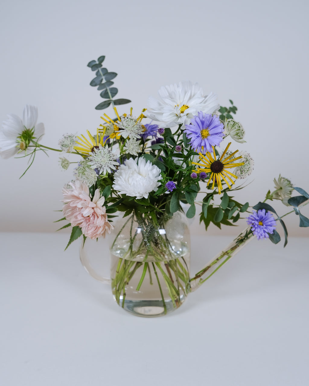 A mixed wildflower bouquet in a clear glass pitcher vase, featuring white daisies, purple flowers, yellow daisy-like blooms, pale pink blossoms, and green foliage on a white surface against a light gray background.