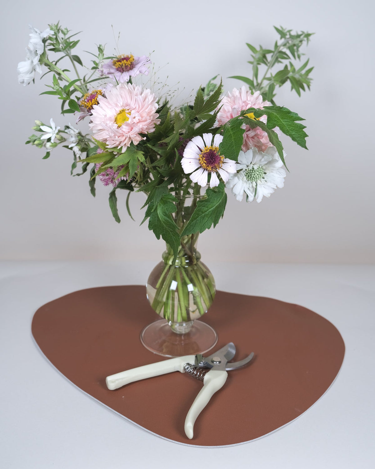 A bouquet of pink, white, and purple wildflowers arranged in a clear glass vase, placed on a brown heart-shaped mat with pruning shears on a light gray background.