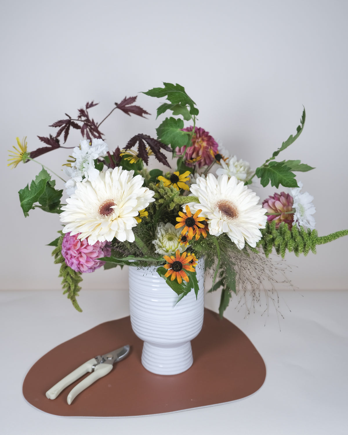 A floral arrangement in a white ribbed vase, featuring white daisies, pink and orange flowers, and green foliage, placed on a brown mat with pruning shears on a light gray background.