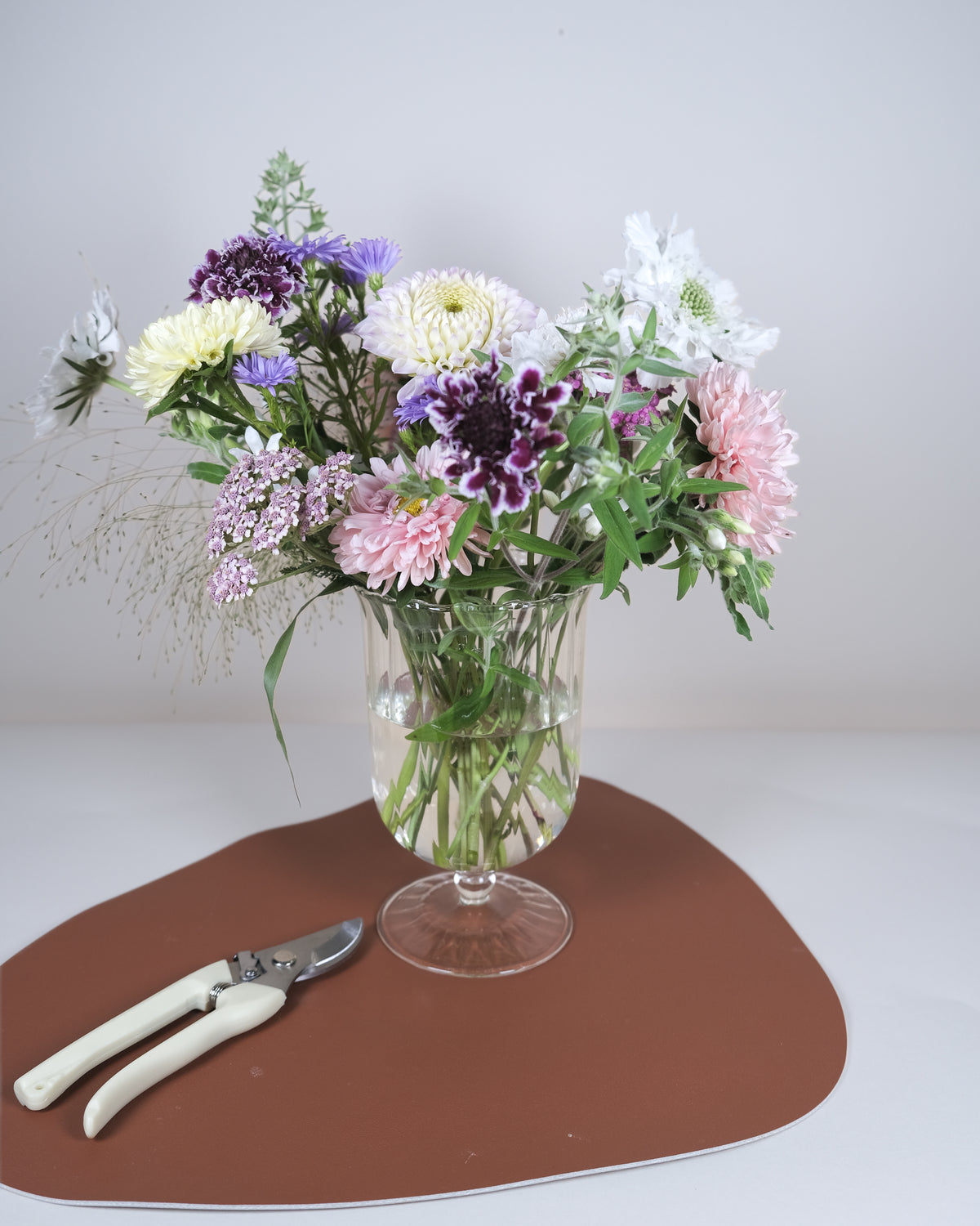 A mixed flower bouquet in a clear glass vase with a stem base, placed on a brown mat with pruning shears nearby against a plain light background.