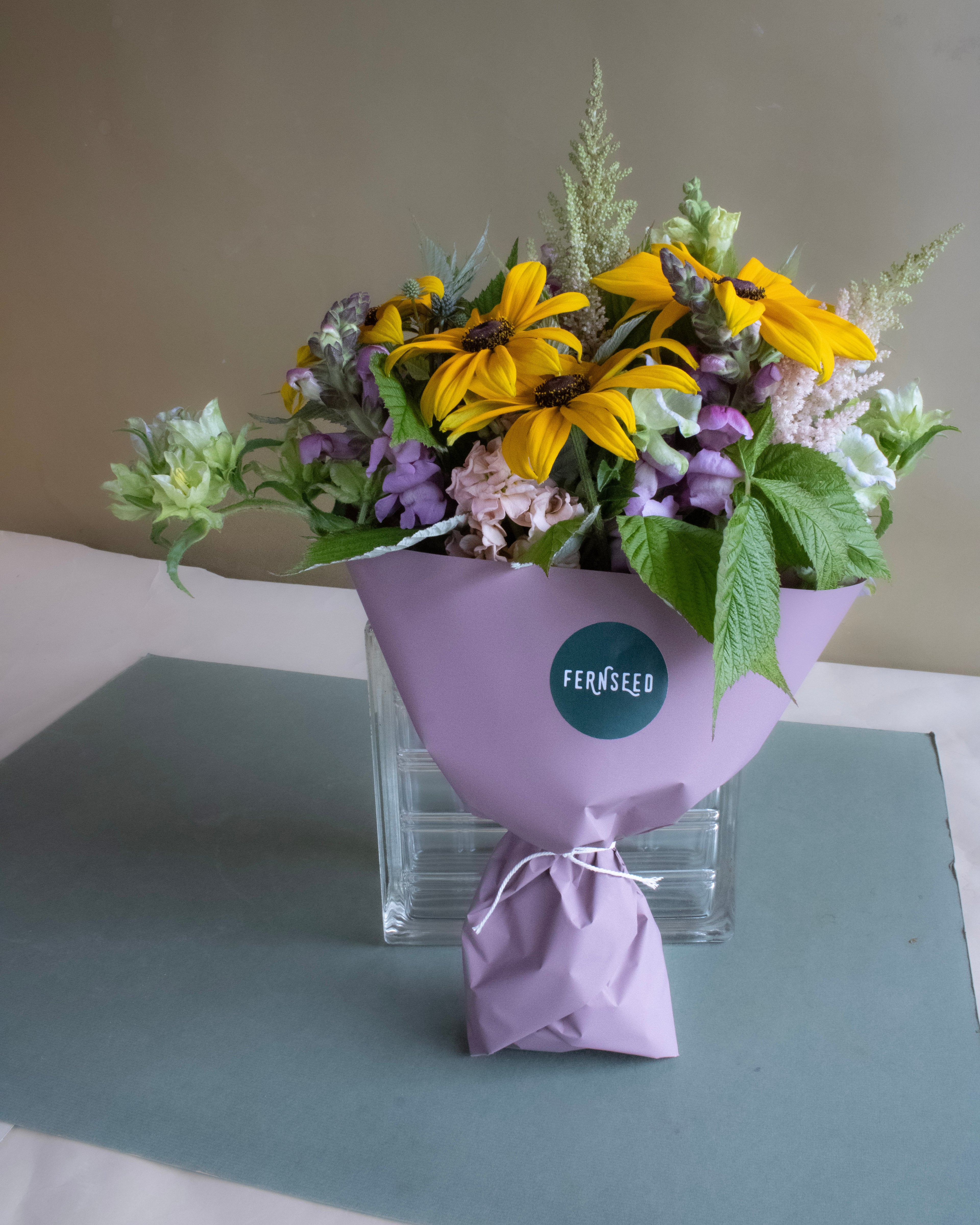 Bouquet of flowers wrapped in purple paper with a Fernseed label on a gray surface.