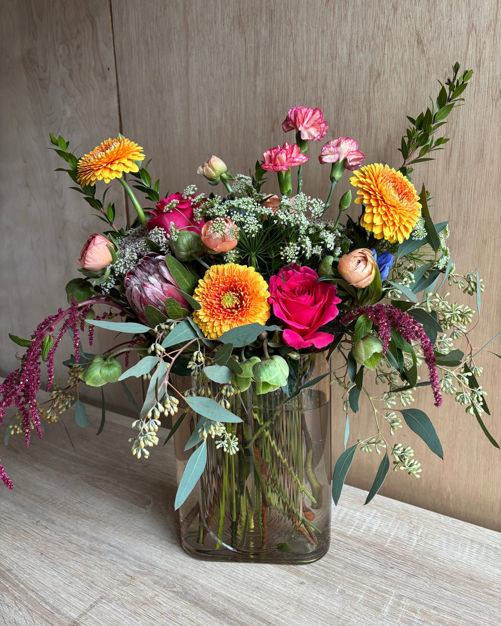 A colorful floral arrangement in a clear glass vase, featuring a variety of flowers with green leaves and stems.
