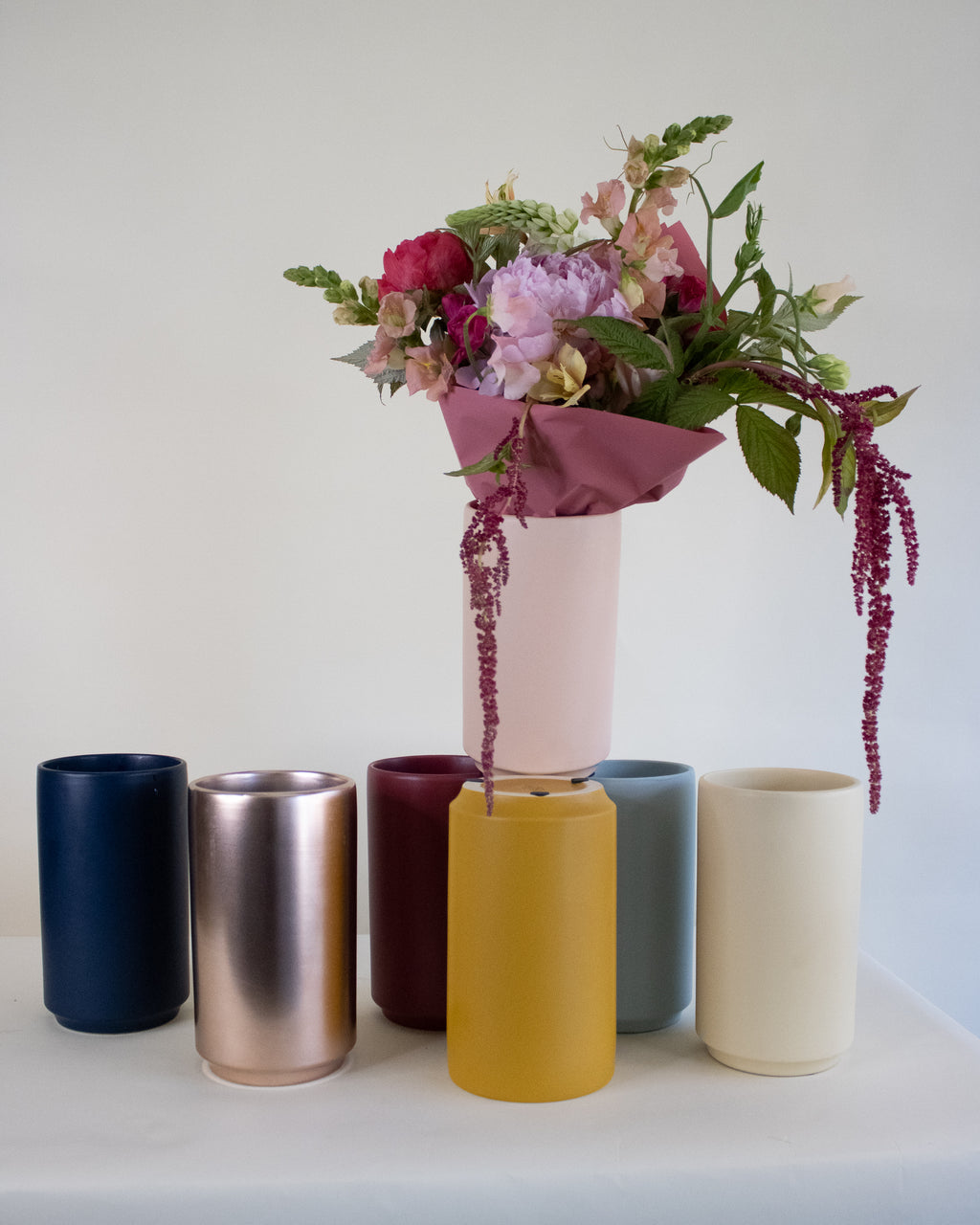Six cylindrical vases in navy, rose gold, burgundy, mustard yellow, slate blue, and cream, arranged against a white backdrop. A blush pink vase stacked on top holds a bouquet of peonies, snapdragons, and trailing burgundy amaranth wrapped in mauve paper.