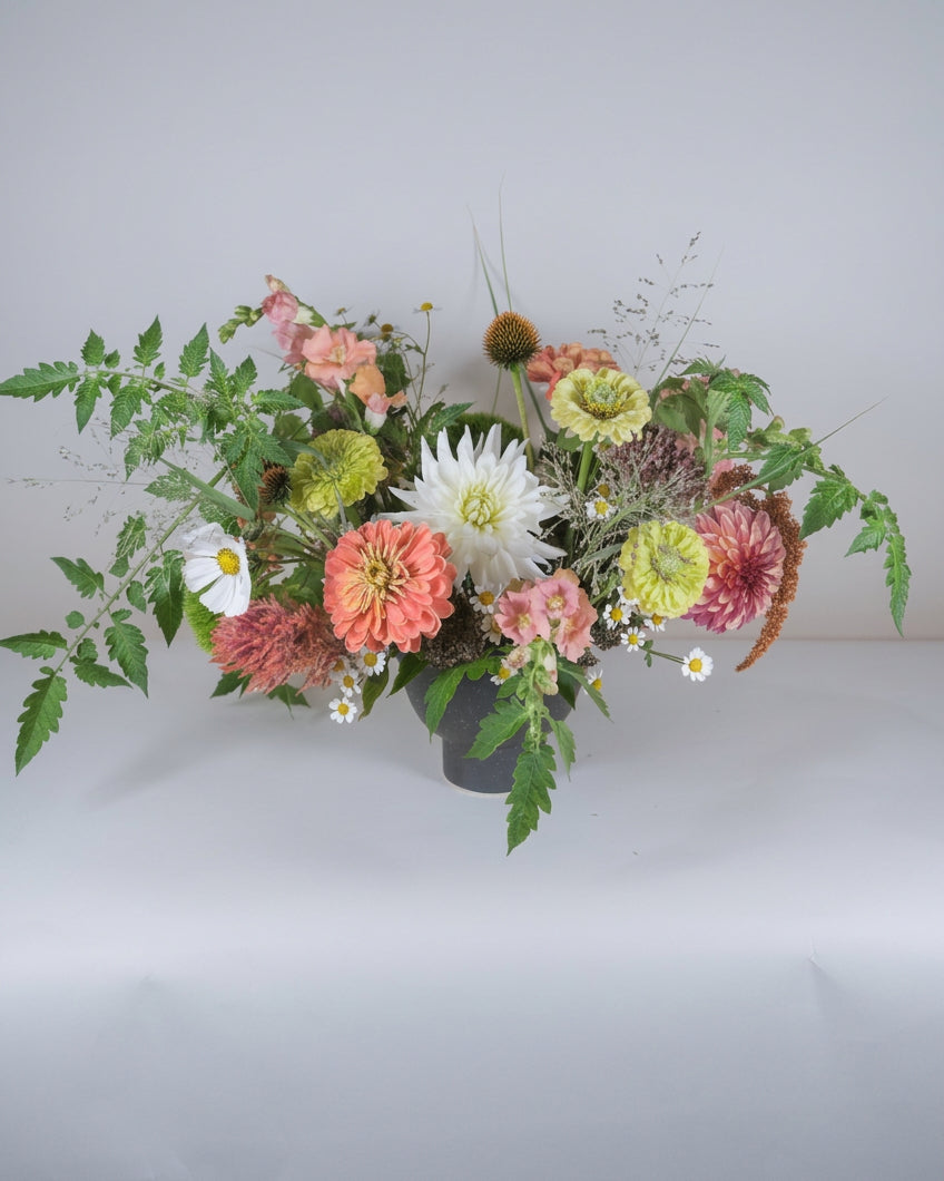 A flower arrangement in a dark gray ceramic pot, featuring white dahlias, pink and orange zinnias, green thistles, and green foliage, set against a plain light gray background.