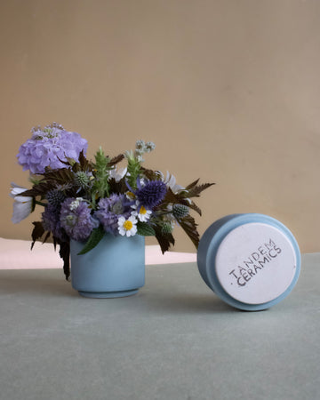 Blue ceramic planter with flowers and lid labeled 'Jewelry Ceramics' on a neutral background