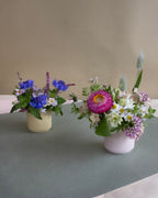 Two flower arrangements in small vases on a gray surface with a beige background
