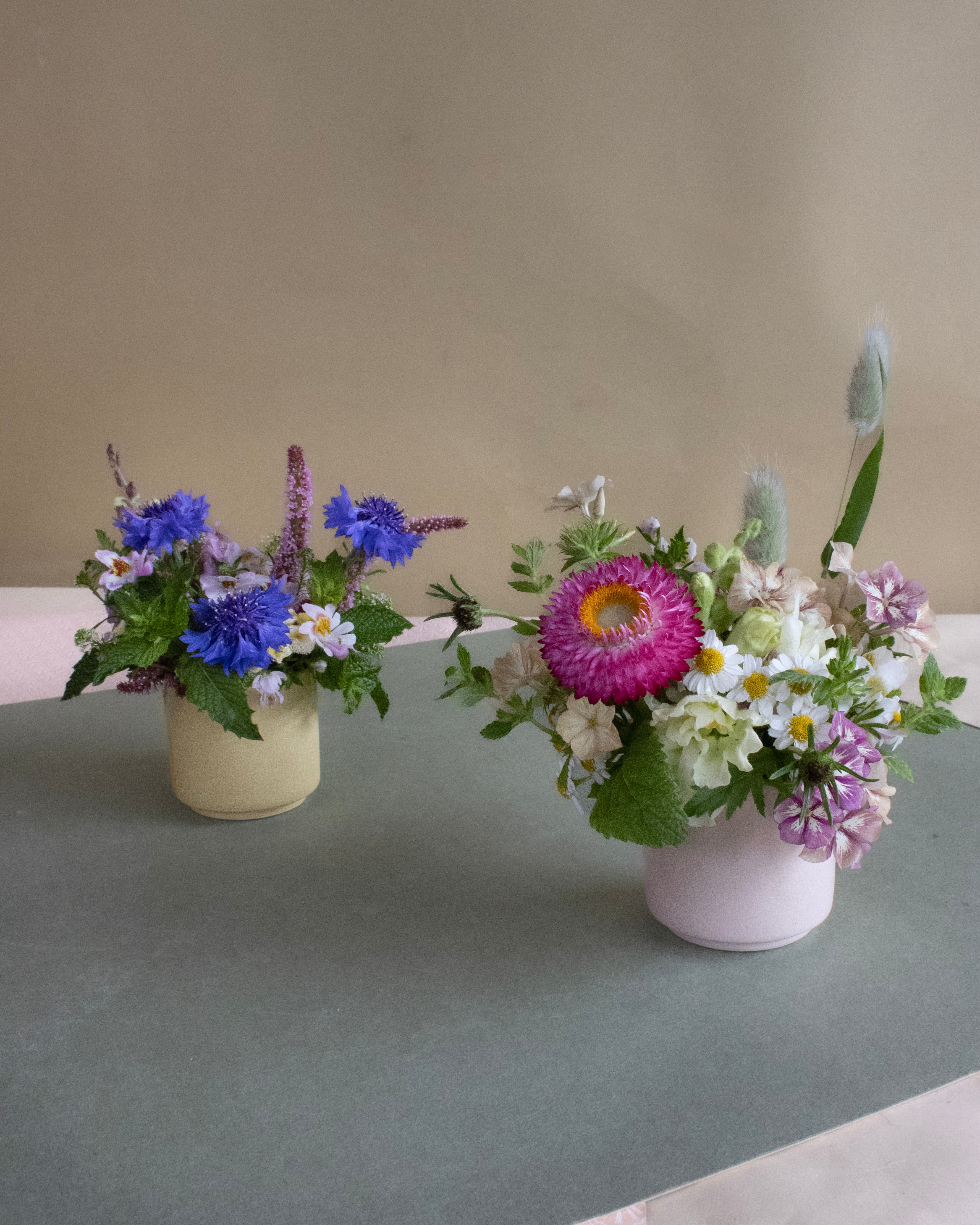 Two flower arrangements in small vases on a gray surface with a beige background