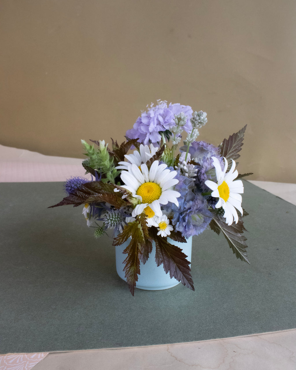 Floral arrangement with daisies and hydrangeas in a white vase on a green surface.