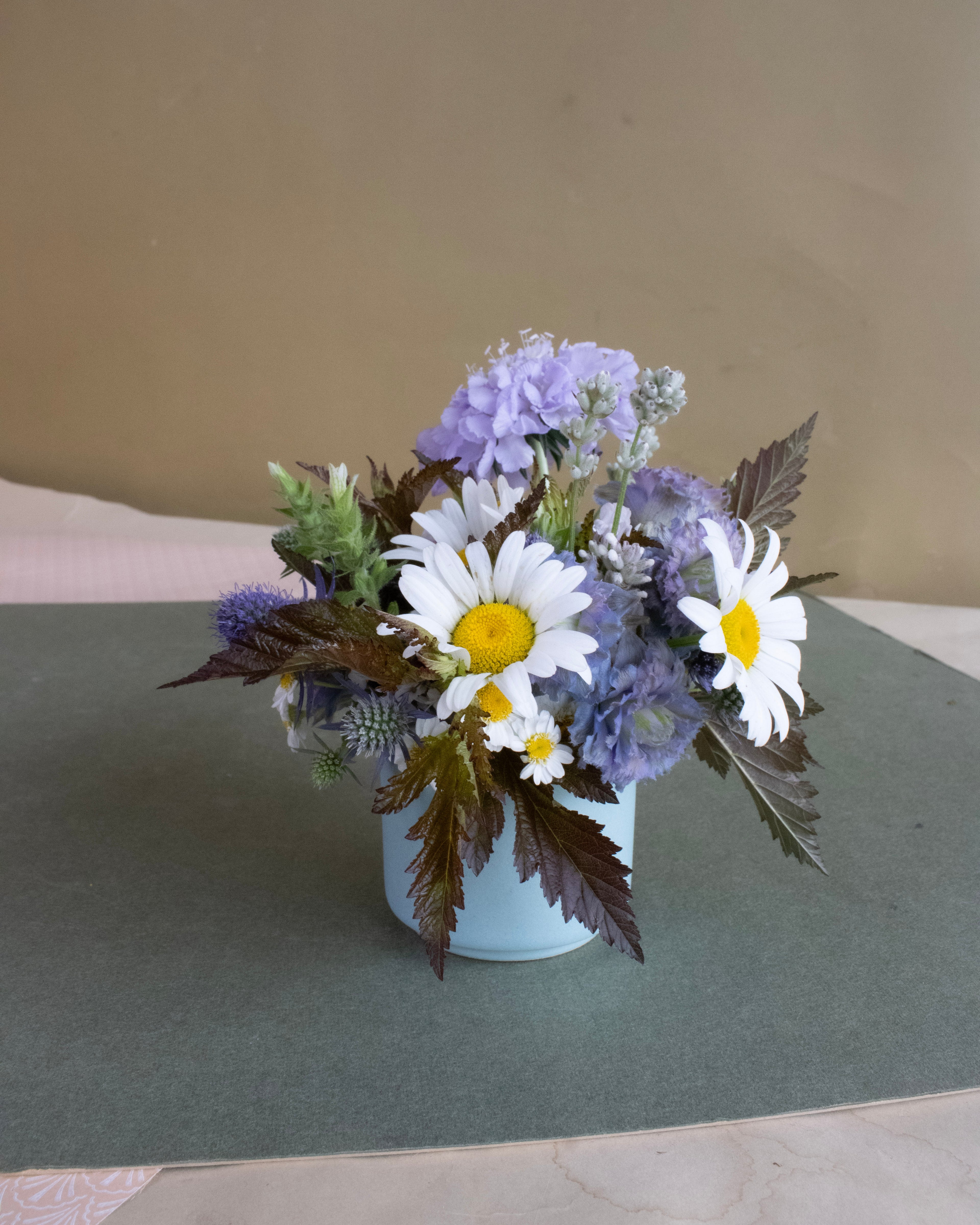Floral arrangement with daisies and hydrangeas in a white vase on a green surface.