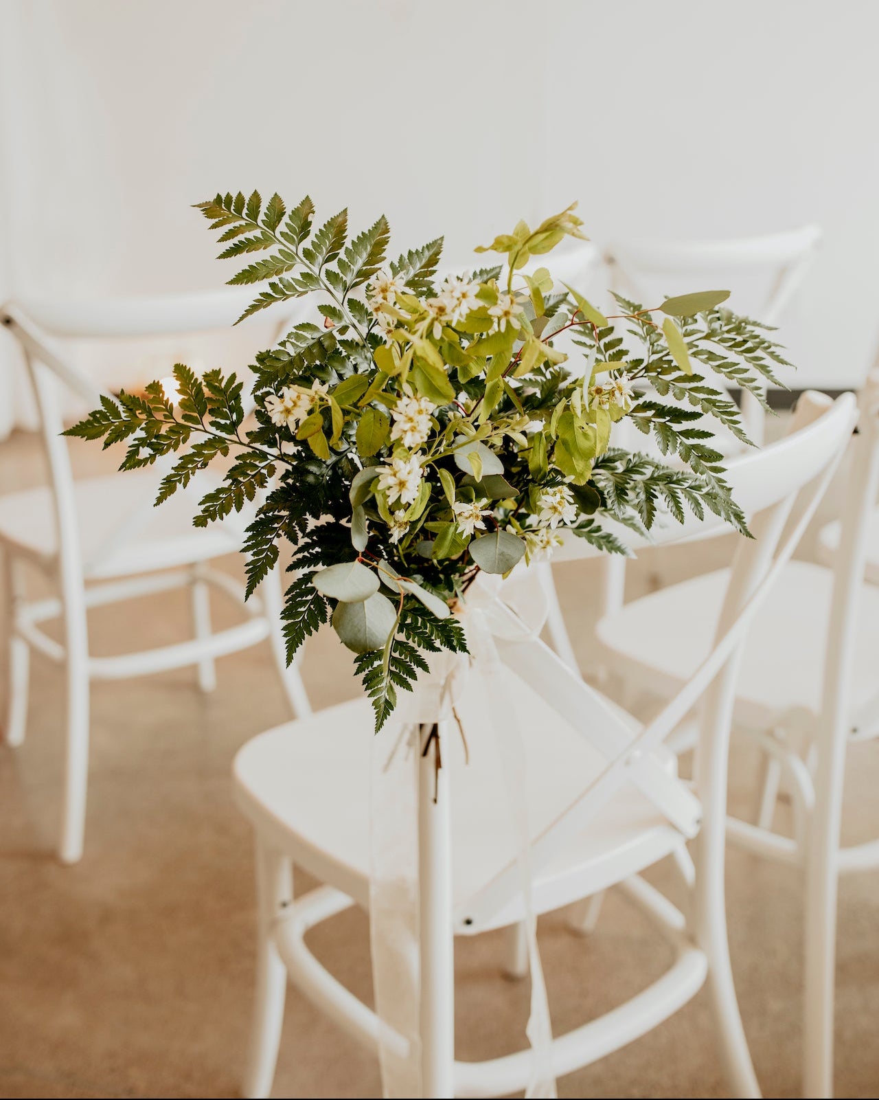 A chair bundle with green foliage and white flowers arranged and tied to a chair, with other chairs in the background.